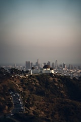 A large observatory with distinctive domes is situated atop a hill, surrounded by greenery. In the background, a sprawling cityscape with numerous tall buildings is visible under a clear sky.