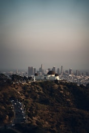A large observatory with distinctive domes is situated atop a hill, surrounded by greenery. In the background, a sprawling cityscape with numerous tall buildings is visible under a clear sky.