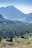 A serene mountain landscape featuring two wooden cabins nestled in a lush green meadow. Dense coniferous forests cover the middle ground, while towering mountain peaks rise impressively in the background under a clear blue sky.