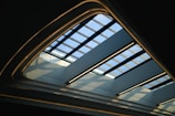 A modern architectural ceiling design featuring a skylight with a grid pattern made of blue-tinted glass panes. The structure is framed with curved, illuminated edges that create a soft glow against the darkened surroundings. Light streams through the glass panes, casting geometric shadows on the interior.