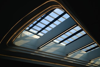 A modern architectural ceiling design featuring a skylight with a grid pattern made of blue-tinted glass panes. The structure is framed with curved, illuminated edges that create a soft glow against the darkened surroundings. Light streams through the glass panes, casting geometric shadows on the interior.