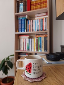 A white mug with a hand-painted watermelon illustration and the phrase 'Say bismillah yallah drink :)' sits on a wooden table. A pink crochet coaster is placed underneath the mug. In the background, there is a wooden bookshelf filled with a variety of colorful books and a small green potted plant is visible.