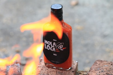 A bottle of hot sauce labeled 'Hola Loca' is placed on bricks with flames in the foreground, creating a dynamic scene.