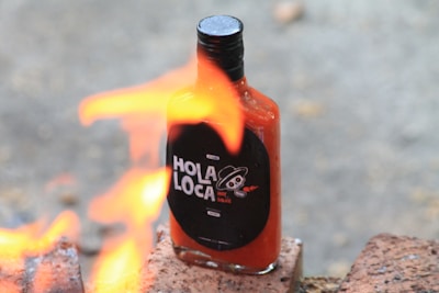 A bottle of hot sauce labeled 'Hola Loca' is placed on bricks with flames in the foreground, creating a dynamic scene.