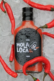 A bottle of hot sauce labeled 'Hola Loca' is surrounded by ice and red chili peppers, with the bottle covered in water droplets. The label features a stylized skull and indicates a best-before date of July 2026.