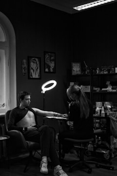 A person is seated in a chair receiving a tattoo on their left arm from a tattoo artist. The room is dimly lit with a ring light focused on the arm. The walls are decorated with artwork and there are shelves filled with various supplies. The setting appears intimate and focused.