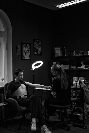 A person is seated in a chair receiving a tattoo on their left arm from a tattoo artist. The room is dimly lit with a ring light focused on the arm. The walls are decorated with artwork and there are shelves filled with various supplies. The setting appears intimate and focused.