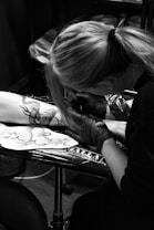 A tattoo artist is working on a client's arm, meticulously inking a detailed dragon design. The artist is wearing gloves and is focused on their work. The setting appears to be a tattoo studio with various equipment visible.