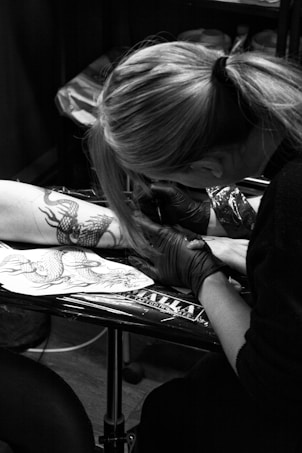 A tattoo artist is working on a client's arm, meticulously inking a detailed dragon design. The artist is wearing gloves and is focused on their work. The setting appears to be a tattoo studio with various equipment visible.