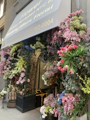 An elaborate floral display surrounds the entrance of a building, featuring a variety of flowers including pink roses, hydrangeas, and other colorful blooms. The entrance is covered by a canopy with the text and phone number of a florist. Greenery and flowers are arranged creatively around door arches and planters, creating a welcoming and vibrant atmosphere.