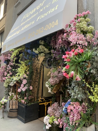 An elaborate floral display surrounds the entrance of a building, featuring a variety of flowers including pink roses, hydrangeas, and other colorful blooms. The entrance is covered by a canopy with the text and phone number of a florist. Greenery and flowers are arranged creatively around door arches and planters, creating a welcoming and vibrant atmosphere.