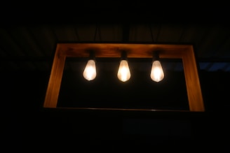 Three light bulbs are mounted in a horizontal wooden frame against a dark background. The bulbs emit a warm glow, creating a cozy atmosphere.