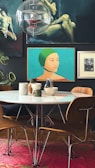 A modern interior featuring a round table surrounded by wooden chairs with a white surface. Several ceramic and stone objects, including vases and bowls, are placed on the table. The background displays a collection of framed artworks on a dark wall, including a portrait of a woman in a green cap. A transparent spherical ceiling light fixture hangs above the table. The floor is covered with a vibrant pink patterned rug, and a large green potted plant is partially visible.