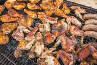 Grilled chicken wings and drumsticks with a charred exterior are arranged on a metal grill, alongside several grilled sausages. The chicken pieces display varying shades of brown and reddish hues, indicating they are well-cooked.