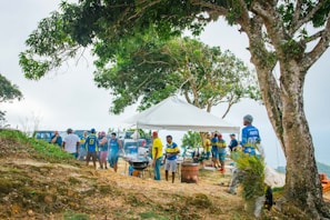 A group of people gathered outdoors under a canopy, surrounded by trees and a hillside. Some individuals are wearing vibrant blue and yellow sports jerseys. Barbecue grills are set up with smoke rising, indicating outdoor cooking activities. The atmosphere suggests a casual gathering or picnic.
