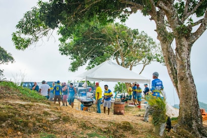 A group of people gathered outdoors under a canopy, surrounded by trees and a hillside. Some individuals are wearing vibrant blue and yellow sports jerseys. Barbecue grills are set up with smoke rising, indicating outdoor cooking activities. The atmosphere suggests a casual gathering or picnic.