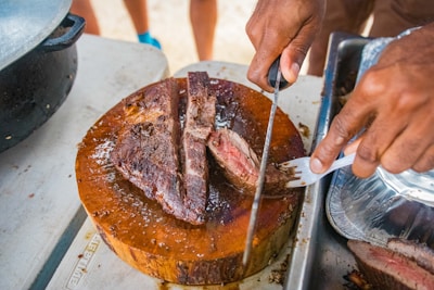 A person is slicing a piece of cooked meat on a round wooden cutting board. The meat appears juicy and cooked to medium-rare, with a pink center visible. The person is using a sharp knife and holding the meat steady with a fork. There is a metal tray beside the cutting board.