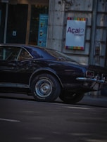 A vintage black car is parked on a city street next to a building with a sign that reads 'Açaí Concept'. The car's shiny surface reflects light, and the building's wall appears to be made of stone. Posters are visible through a nearby glass door.