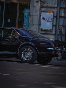 A vintage black car is parked on a city street next to a building with a sign that reads 'Açaí Concept'. The car's shiny surface reflects light, and the building's wall appears to be made of stone. Posters are visible through a nearby glass door.