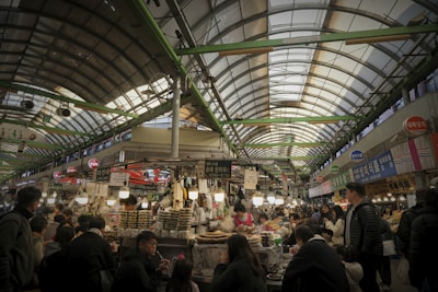 A bustling indoor market scene with numerous people shopping and eating at various food stalls. The market is covered with a high arched roof supported by green beams. Vendors display their goods under illuminated signs, and the atmosphere is lively with a mixture of transactions and conversations among shoppers.