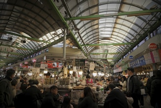 A bustling indoor market scene with numerous people shopping and eating at various food stalls. The market is covered with a high arched roof supported by green beams. Vendors display their goods under illuminated signs, and the atmosphere is lively with a mixture of transactions and conversations among shoppers.