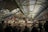 A bustling indoor market scene with numerous people shopping and eating at various food stalls. The market is covered with a high arched roof supported by green beams. Vendors display their goods under illuminated signs, and the atmosphere is lively with a mixture of transactions and conversations among shoppers.