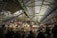A bustling indoor market scene with numerous people shopping and eating at various food stalls. The market is covered with a high arched roof supported by green beams. Vendors display their goods under illuminated signs, and the atmosphere is lively with a mixture of transactions and conversations among shoppers.