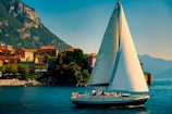 A sailboat with a large white sail glides across a serene body of water. In the background, quaint houses with terracotta roofs line the waterfront, nestled against a lush green mountainside. The sky is clear with a soft blue hue, and people can be seen on the boat, enjoying leisure time.