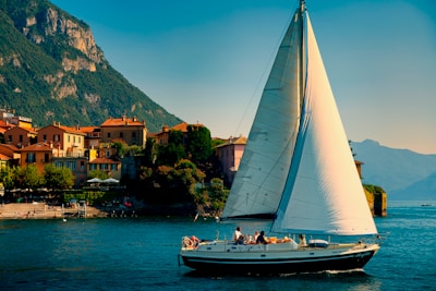 A sailboat with a large white sail glides across a serene body of water. In the background, quaint houses with terracotta roofs line the waterfront, nestled against a lush green mountainside. The sky is clear with a soft blue hue, and people can be seen on the boat, enjoying leisure time.