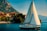A sailboat with a large white sail glides across a serene body of water. In the background, quaint houses with terracotta roofs line the waterfront, nestled against a lush green mountainside. The sky is clear with a soft blue hue, and people can be seen on the boat, enjoying leisure time.