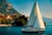 A sailboat with a large white sail glides across a serene body of water. In the background, quaint houses with terracotta roofs line the waterfront, nestled against a lush green mountainside. The sky is clear with a soft blue hue, and people can be seen on the boat, enjoying leisure time.