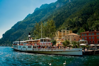 A vintage-style passenger boat is docked at a lakeside town surrounded by lush green mountains. The boat is filled with people enjoying the scenic view. The architecture of the town features colorful buildings with red, yellow, and cream facades. A few swans are visible in the water near the boat.