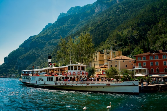A vintage-style passenger boat is docked at a lakeside town surrounded by lush green mountains. The boat is filled with people enjoying the scenic view. The architecture of the town features colorful buildings with red, yellow, and cream facades. A few swans are visible in the water near the boat.