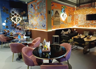 A cozy indoor cafe setting featuring round black tables paired with plush pink chairs and dark, geometric-patterned sofas. The walls are adorned with vibrant, eclectic art pieces, mixing abstract and graffiti elements. Illuminated signs and a warm lighting scheme create an inviting atmosphere, highlighting shelves with decor items and bottled water.