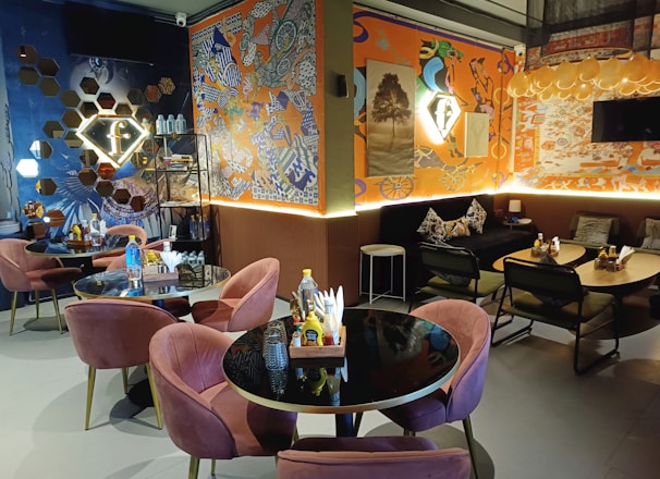 A cozy indoor cafe setting featuring round black tables paired with plush pink chairs and dark, geometric-patterned sofas. The walls are adorned with vibrant, eclectic art pieces, mixing abstract and graffiti elements. Illuminated signs and a warm lighting scheme create an inviting atmosphere, highlighting shelves with decor items and bottled water.