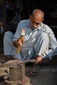 An older man is sitting on the ground while working with a hammer on a piece of metal placed on an anvil. He is wearing light-colored traditional clothing and sandals. There is a watch on his left wrist. The background includes various workshop tools and materials, partially in shadow.