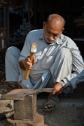 An older man is sitting on the ground while working with a hammer on a piece of metal placed on an anvil. He is wearing light-colored traditional clothing and sandals. There is a watch on his left wrist. The background includes various workshop tools and materials, partially in shadow.