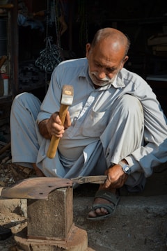 An older man is sitting on the ground while working with a hammer on a piece of metal placed on an anvil. He is wearing light-colored traditional clothing and sandals. There is a watch on his left wrist. The background includes various workshop tools and materials, partially in shadow.