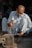 An older man is sitting on the ground while working with a hammer on a piece of metal placed on an anvil. He is wearing light-colored traditional clothing and sandals. There is a watch on his left wrist. The background includes various workshop tools and materials, partially in shadow.