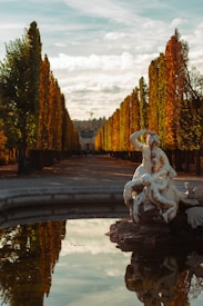 A marble statue depicting a mythological scene is situated at the center of a reflecting pool. The background features a symmetrical row of tall trees with autumn foliage, leading the eye towards a distant building. The sky is partly cloudy, casting a warm, golden light over the scene.