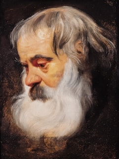 An elderly man's profile with a full white beard and thinning, long white hair. His skin tone is warm, and the expression appears contemplative or somber. The background is dark, highlighting the features of the face.