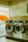 A row of industrial washing machines with a capacity of 7 kg is lined up in a laundromat. The machines are metallic and numbered, with doors that are slightly ajar. Light from outside comes through a partially visible window featuring text in orange.