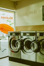 A row of industrial washing machines with a capacity of 7 kg is lined up in a laundromat. The machines are metallic and numbered, with doors that are slightly ajar. Light from outside comes through a partially visible window featuring text in orange.
