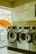 A row of industrial washing machines with a capacity of 7 kg is lined up in a laundromat. The machines are metallic and numbered, with doors that are slightly ajar. Light from outside comes through a partially visible window featuring text in orange.