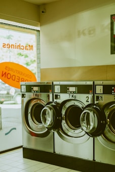 A row of industrial washing machines with a capacity of 7 kg is lined up in a laundromat. The machines are metallic and numbered, with doors that are slightly ajar. Light from outside comes through a partially visible window featuring text in orange.
