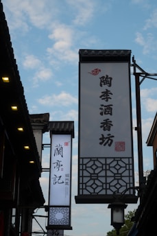Several traditional signs with Chinese characters hang prominently against a backdrop of a partly cloudy sky. The signs are affixed to a building with intricate architectural details, including tiled roofs and lantern-style lighting.
