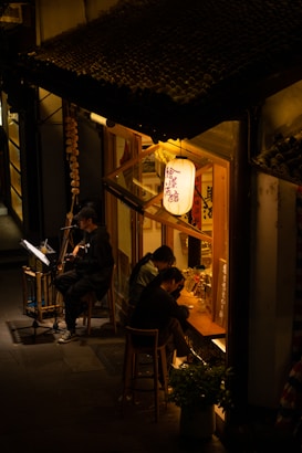 A cozy street scene at night featuring a small, warmly lit restaurant or bar with traditional lanterns. Two people are sitting at the counter inside, and a musician is playing guitar outside, illuminated by a light. There are decorative elements such as strings of beads and plants adorning the exterior.