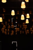 Numerous hanging lamps cast a warm glow in a dimly lit room, creating a serene and slightly mysterious atmosphere. The lamps are uniformly arranged and contribute soft lighting to the dark surroundings.