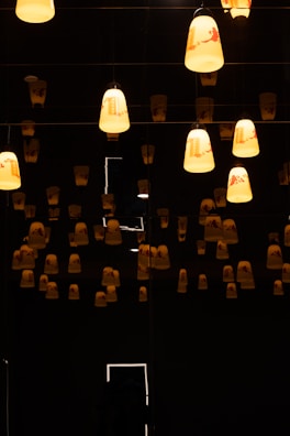 Numerous hanging lamps cast a warm glow in a dimly lit room, creating a serene and slightly mysterious atmosphere. The lamps are uniformly arranged and contribute soft lighting to the dark surroundings.