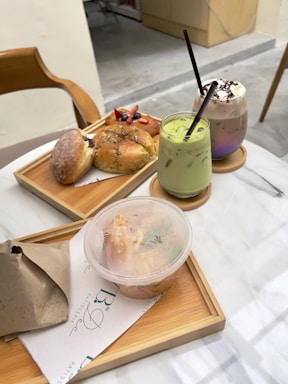 A table setting displaying a selection of pastries and beverages. There are a couple of pastries on a wooden tray, one topped with fruit and another with herbs. Next to them are two drinks in glasses, one with a green liquid appearing to be matcha and another topped with whipped cream and chocolate syrup. There is also a plastic container with salad and a wrapped item on the tray.